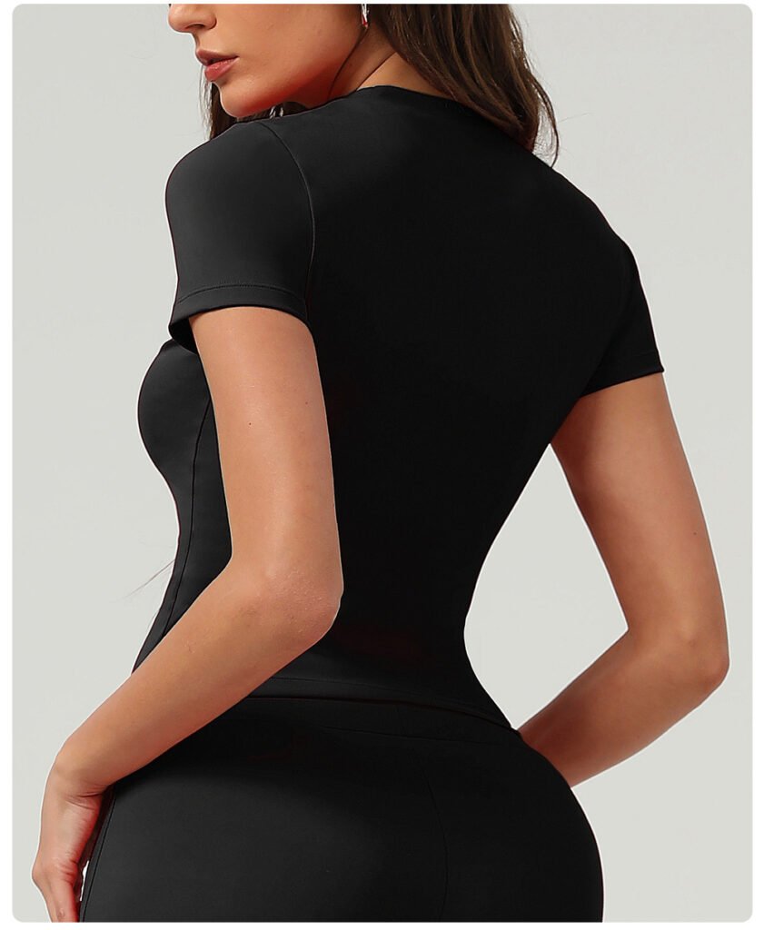 Back angle close-up black stretch workout tee, vertical waist contour seaming, seamless short sleeves matte sports fabric neutral wall background