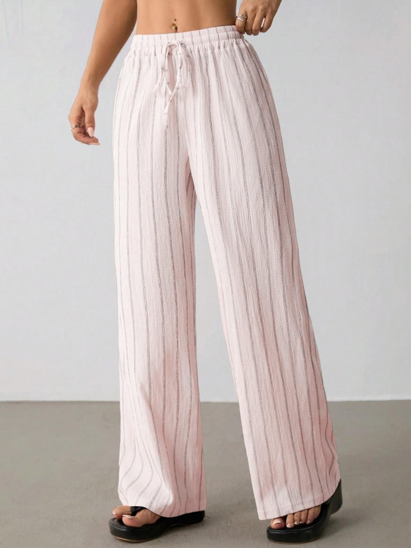 Pastel Pink Striped Wide Leg Pants Drawstring Elastic Waist Crinkle Fabric Side angle pastel pink vertical stripe palazzo pants, full ruffle elastic waist, matching stripe drawstring tie, lightweight crinkle textured woven fabric indoor partial outfit photo